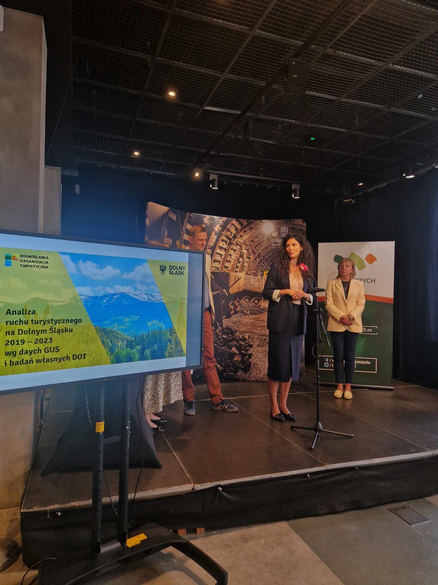 Zdjęcie przedstawia wysoką kobietę zapowiadającą imprezę podczas konferencji w oświetlonym pomieszczeniu