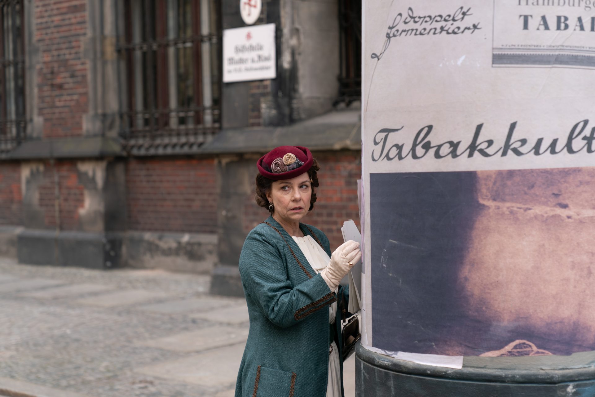 Kobieta w stroju z lat 30-tych stoi przy słupie reklamowym.